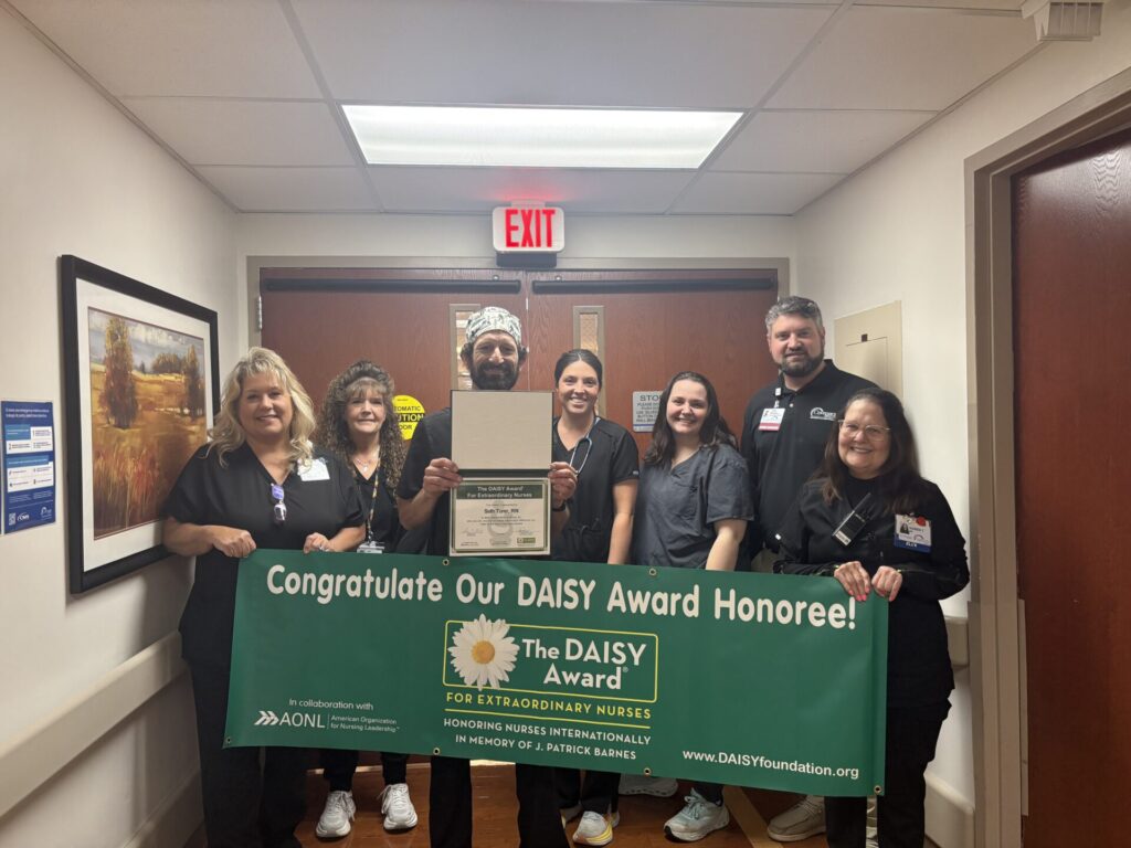 Seven people stand in a hallway, smiling and holding a green banner that reads, Congratulate Our DAISY Award Honoree! One person in the middle holds a certificate and a small statue.