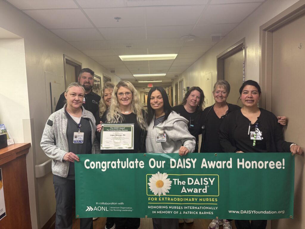 A group of nurses stand in a hallway holding a green banner that reads Congratulate Our DAISY Award Honoree! One nurse in the center holds a certificate, and everyone is smiling at the camera.