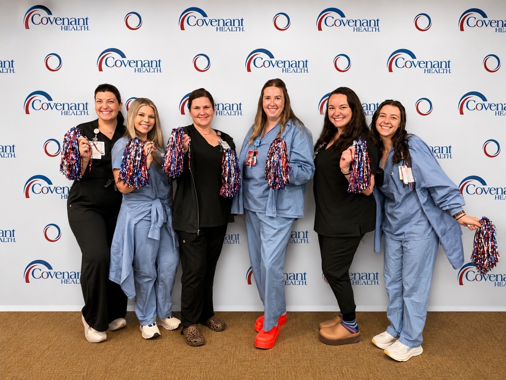 Six smiling healthcare workers pose in front of a Covenant Health backdrop, holding red, white, and blue pom-poms. Some wear scrubs and others wear black uniforms. They stand close together on a tan carpeted floor.