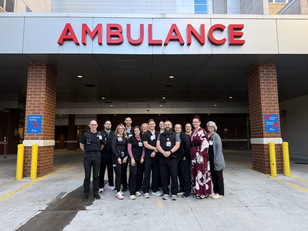 A group of people in medical uniforms stands together smiling outside a hospital emergency entrance beneath a large red AMBULANCE sign. Two people in civilian clothes stand at each end of the group.