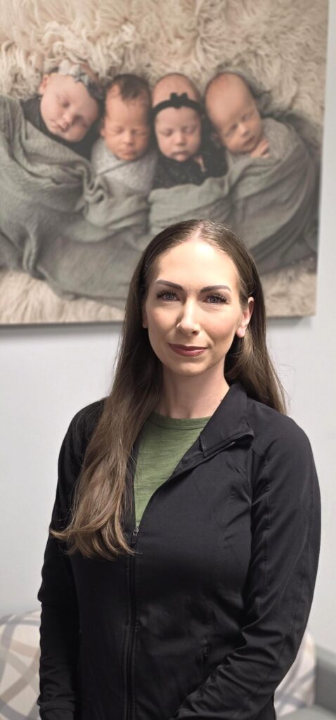 A woman with long brown hair, wearing a black jacket over a green top, stands indoors. Behind her is a framed photo of four swaddled babies lying side by side on a fluffy blanket.