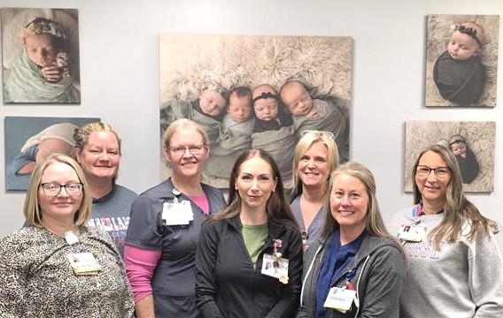 Seven women, some in medical uniforms with badges, stand smiling in front of a wall decorated with photos of newborn babies. The setting appears to be a hospital or medical office.