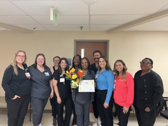 A group of eleven smiling people stand indoors; the woman in the center holds a bouquet of flowers and a certificate, suggesting a celebration or recognition event.