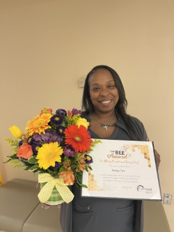 A smiling woman holds a colorful bouquet of flowers and a framed certificate that reads BEE Award in a beige room.