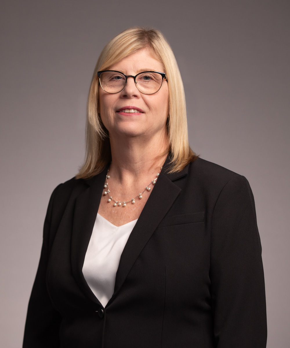 A middle-aged woman with blonde hair wearing glasses, a black blazer, a white blouse, and a pearl necklace stands against a plain, light gray background, looking at the camera with a neutral expression.