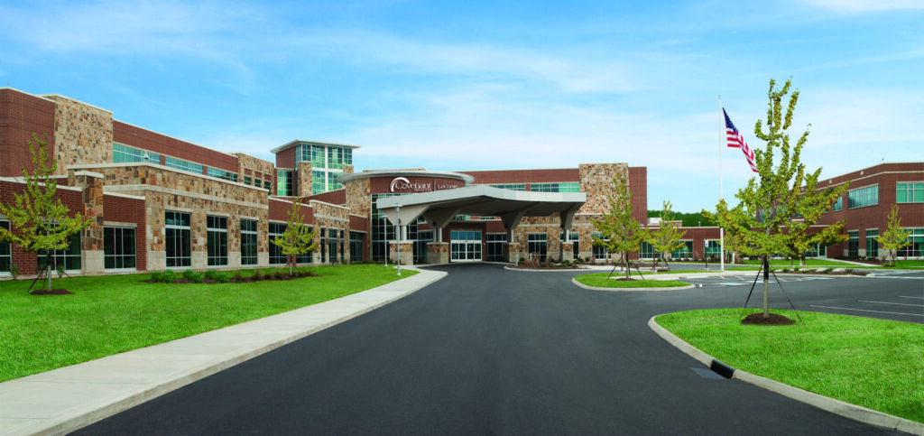 A modern brick and stone hospital building with large windows, an arched entrance, and a covered drop-off area. An American flag flies on the right, with green lawns and trees lining the curved driveway.