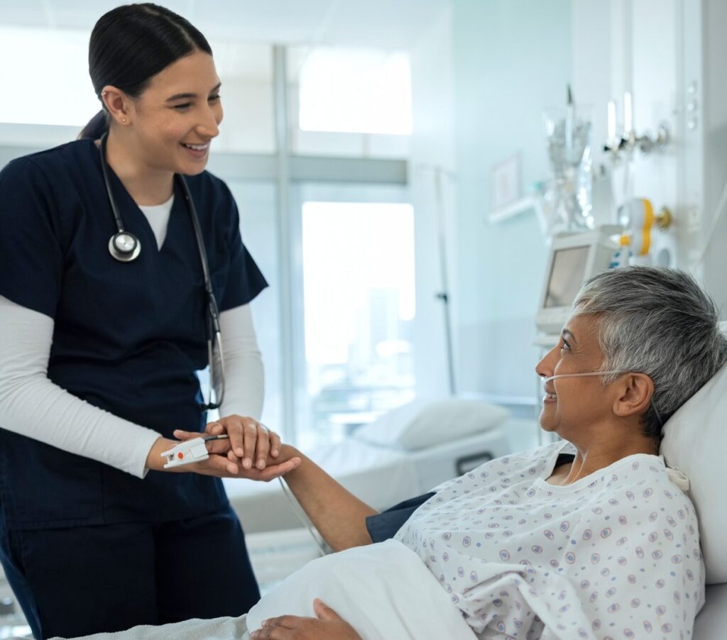 A nurse in navy scrubs smiles and holds the hand of an older patient lying in a hospital bed with an oxygen tube, creating a warm and caring atmosphere in a bright hospital room.