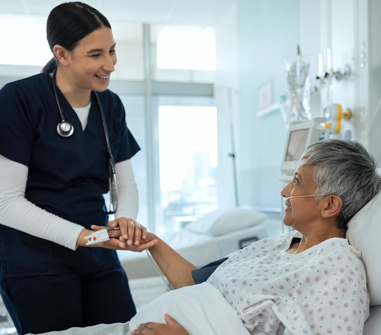 A nurse in navy scrubs smiles and holds the hand of an older patient lying in a hospital bed with an oxygen tube, creating a warm and caring atmosphere in a bright hospital room.