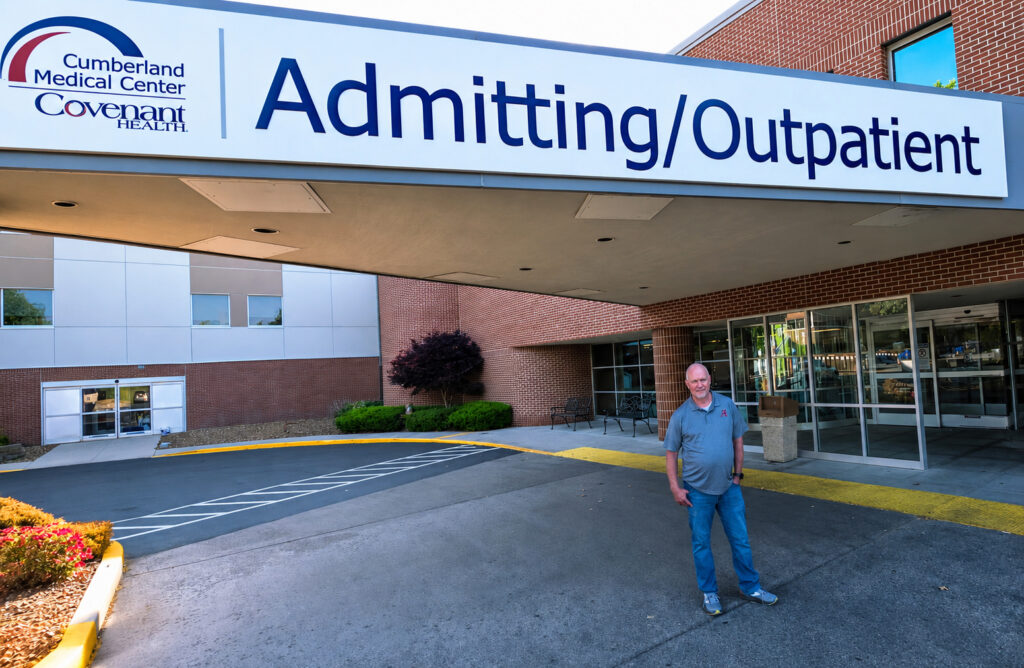 A man stands outside the entrance of the Admitting/Outpatient area at Cumberland Medical Center, part of Covenant Health. The building has large windows and brick walls.