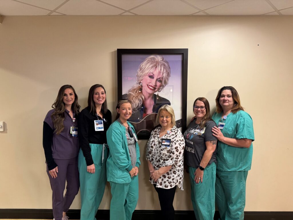 Six women in medical scrubs and badges stand smiling in front of a framed photo of Dolly Parton on a beige wall. They appear to be hospital staff posing together.