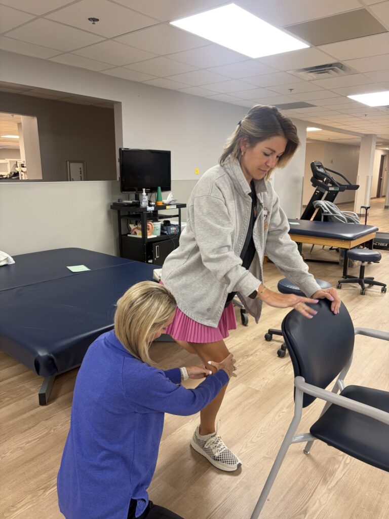A physical therapist kneels beside a woman in athletic clothing, assisting her as she performs a standing exercise using a chair for support in a rehabilitation facility.