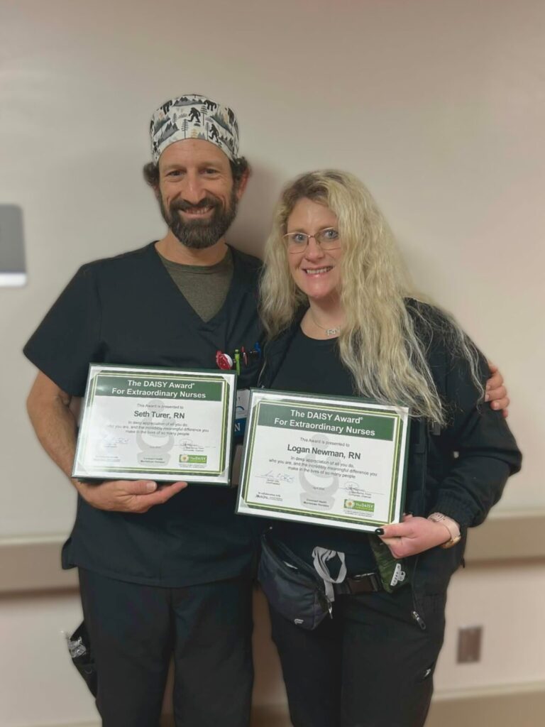 Two nurses in scrubs smile while holding framed certificates for the DAISY Award for Extraordinary Nurses. Both stand side by side in a hospital setting, celebrating their achievement.