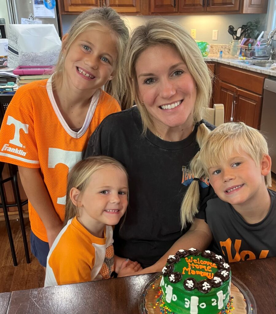 A woman smiles with three young children around a table, celebrating with a small football-themed cake that reads Welcome Home Mommy. They are in a cozy kitchen setting.