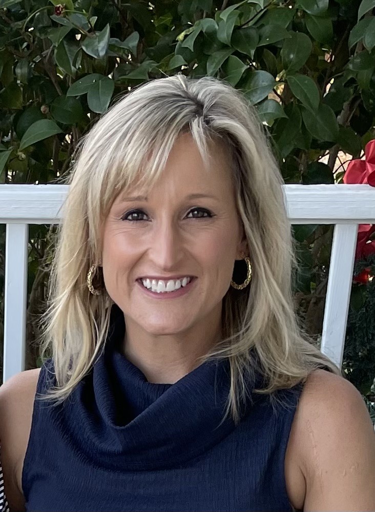 A woman with blonde hair and hoop earrings smiles while sitting in front of green leafy plants and a white fence, wearing a sleeveless navy blue top.