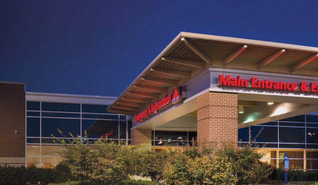 Exterior view of Fort Loudoun Medical Center at dusk, showing the main entrance with a red sign and illuminated hospital logo on a brick wall, surrounded by trees and bushes.