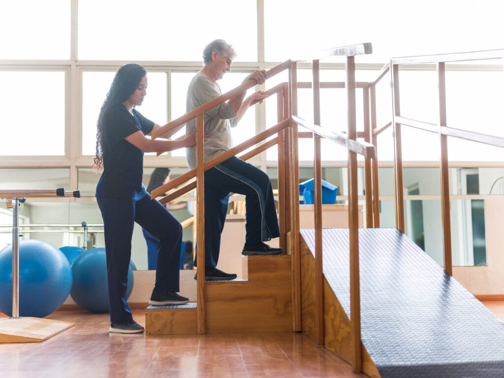 An older adult man practices walking up wooden stairs with handrails, assisted by a female physical therapist in a rehabilitation center with large windows and exercise equipment visible in the background.