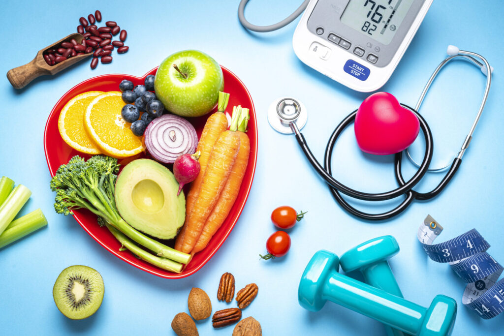 A heart-shaped plate with fruits and vegetables, surrounded by a stethoscope, blood pressure monitor, blue dumbbells, measuring tape, nuts, and beans, symbolizing healthy lifestyle and heart health on a blue background.