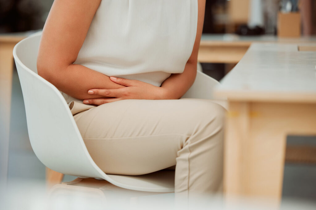 A person sitting in a chair indoors, wearing a sleeveless white top and beige pants, with one hand resting on their stomach, appearing to experience discomfort or pain.