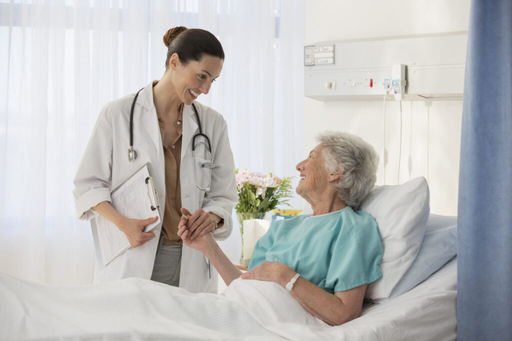 A female doctor smiles and holds the hand of an elderly woman who is lying in a hospital bed, both appearing engaged and positive in a bright, clean hospital room.