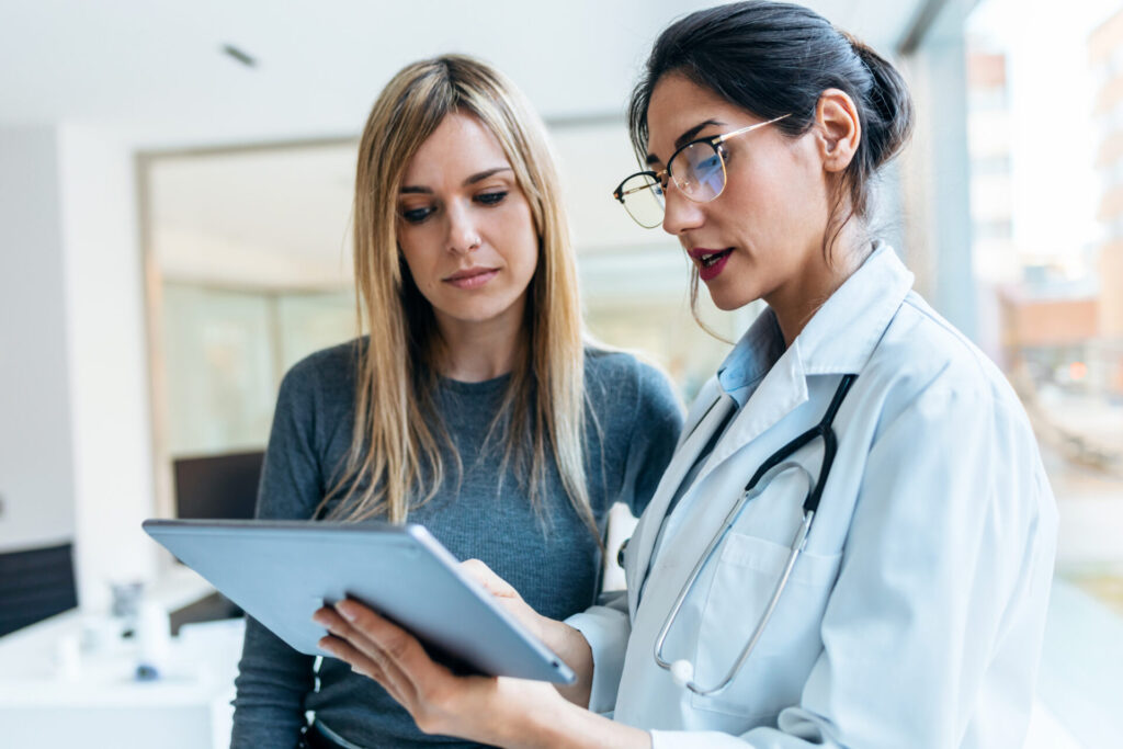 A doctor wearing a white coat and stethoscope shows a tablet to a woman in casual clothes. They stand together indoors, discussing information about autoimmune disease displayed on the screen.
