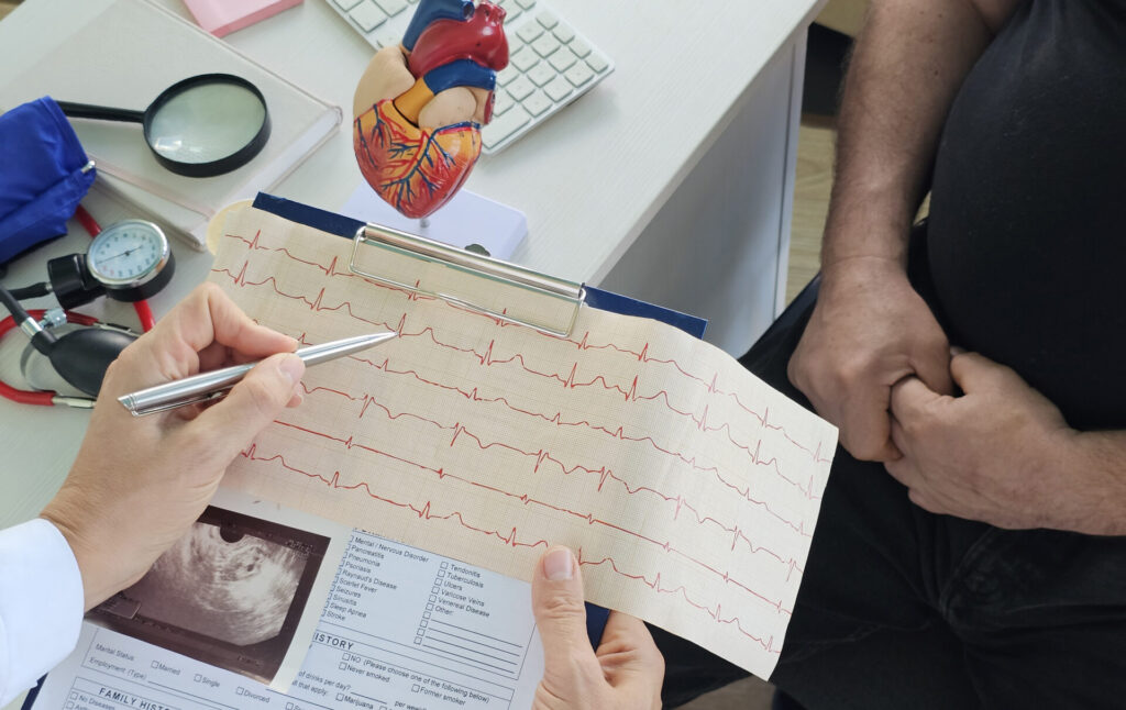 A doctor shows a patient an electrocardiogram (ECG) printout on a clipboard. A heart model, ultrasound image, and medical documents are on the desk nearby. The patient sits with hands folded.