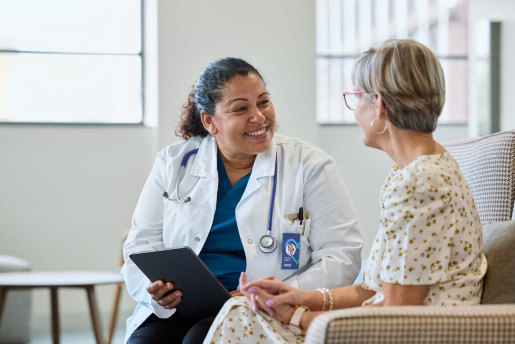 A female doctor smiles while holding a tablet and talking with a seated older woman in a light, modern medical office. The two appear engaged in a friendly, supportive conversation.
