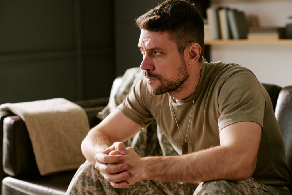 A man in military uniform sits on a couch indoors, leaning forward with his hands clasped, looking thoughtful and serious. Dog tags hang from his neck and books are visible in the background.