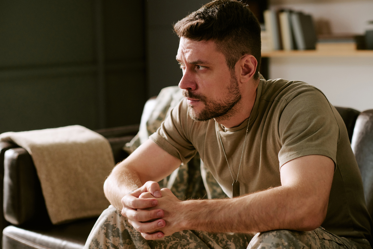A man in military uniform sits on a couch indoors, leaning forward with his hands clasped, looking thoughtful and serious. Dog tags hang from his neck and books are visible in the background.