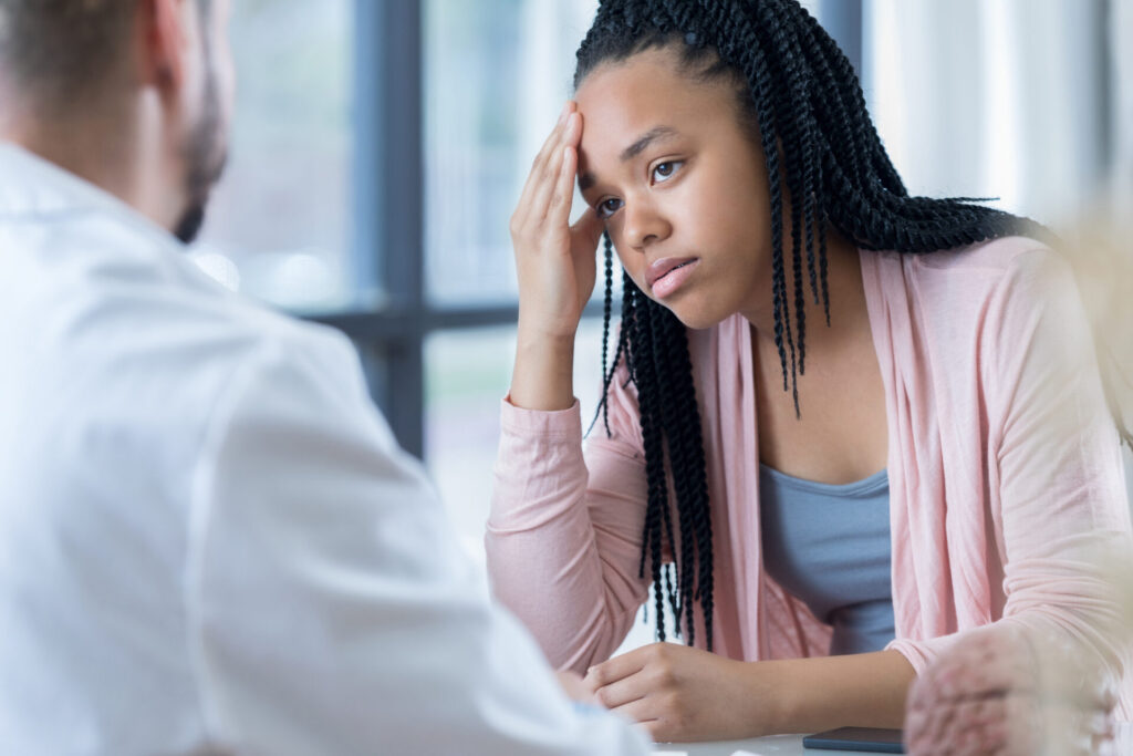 A young woman with braided hair looks concerned, resting her head on her hand while talking to a man in a white coat, possibly a doctor, in a bright office setting.