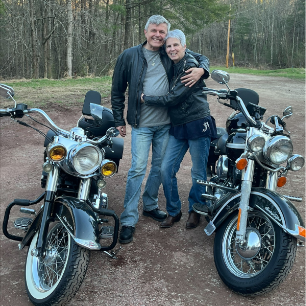 Two people stand smiling with arms around each other between two parked motorcycles on a dirt road, surrounded by trees and greenery in the background.