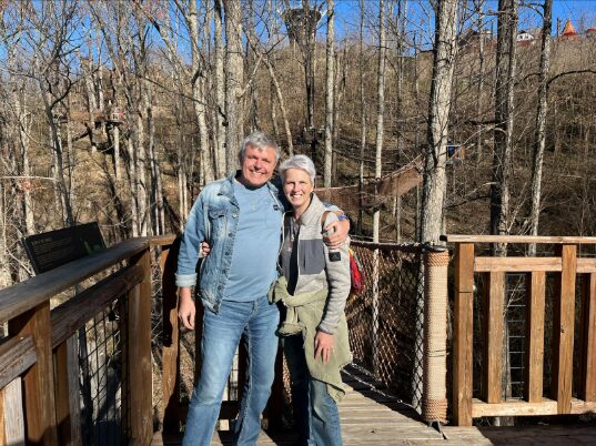 A smiling couple stands on a wooden platform surrounded by leafless trees on a sunny day, with a blue sky and forested hills in the background.