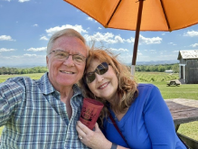 An older couple smiles and poses for a selfie outdoors under an orange umbrella, with green fields, a farmhouse, and blue sky with clouds in the background. The woman holds a drink and both look happy and relaxed.