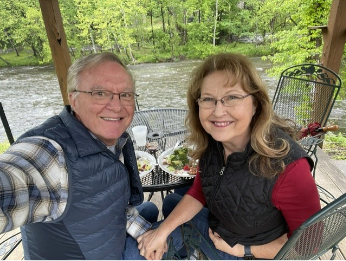 Smiling older couple sitting at an outdoor table by a river, enjoying a meal together under a wooden gazebo, surrounded by lush green trees.
