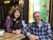 A woman and a man, both wearing plaid shirts, smile while sitting at a wooden counter in what appears to be a cozy café or outdoor market setting.