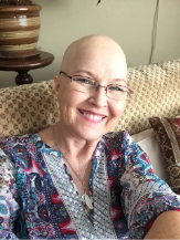 A smiling woman with glasses and a patterned top sits on a couch. She has a bald head and is taking a selfie in a cozy living room.