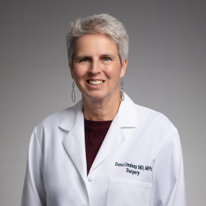 A smiling woman with short gray hair wearing a white lab coat, maroon top, and dangling earrings stands against a plain gray background. Her lab coat reads, &ldquo;Surgery.&rdquo;.