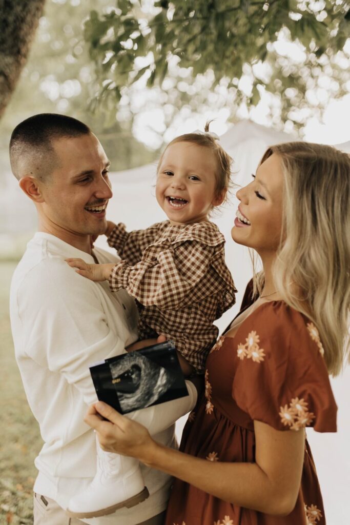 A smiling man and woman hold a happy toddler and an ultrasound photo outdoors under a tree, suggesting they are expecting another child. The family looks joyful and excited together.