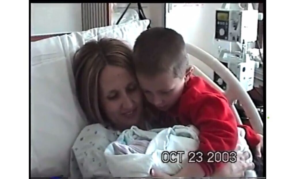 A woman in a hospital bed holds a newborn baby wrapped in a blanket while a young boy in a red shirt hugs them both. 