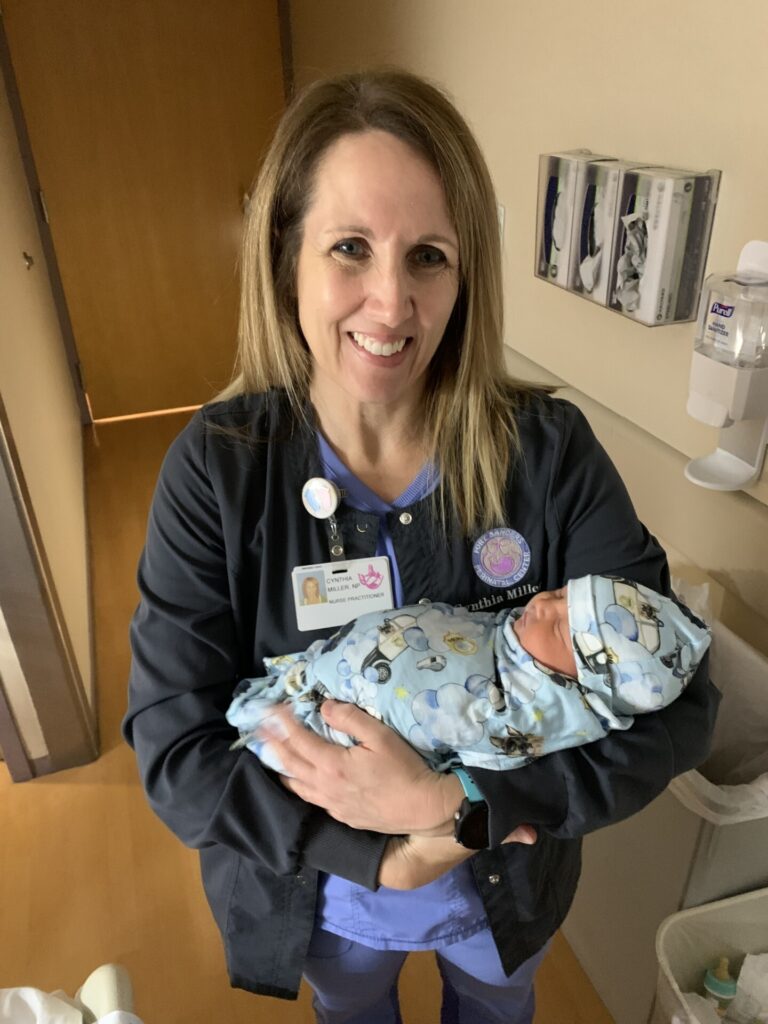 A smiling nurse in scrubs holds a newborn baby wrapped in a blue patterned blanket and hat inside a hospital room. Medical supplies are visible on the wall behind them.