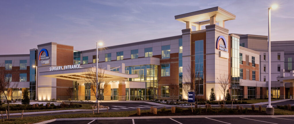 A modern hospital building at dusk features large windows, multiple stories, and a well-lit entrance labeled Surgery Entrance. The surroundings include landscaped grounds and an empty parking lot in the foreground.