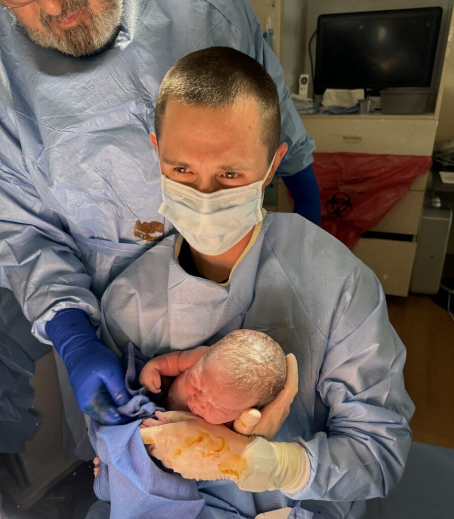 A masked person in medical scrubs holds a newborn baby, also covered in birth fluids, in a hospital setting. Another gloved individual is assisting nearby.