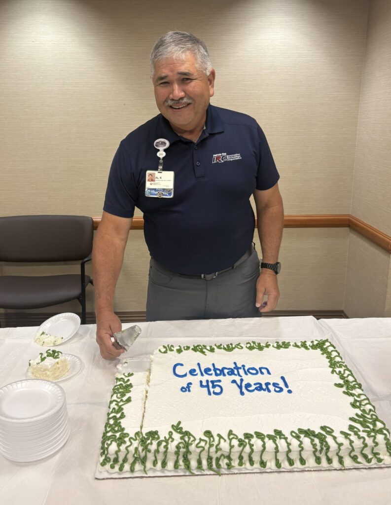A smiling man stands behind a large cake that reads Celebration of 45 Years! as he prepares to serve a slice. He wears a navy shirt with a name badge and stands in a room with beige walls and chairs.