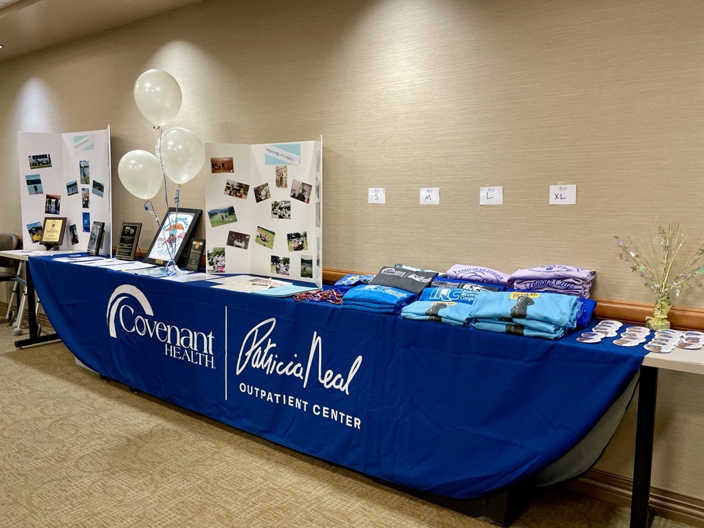 A display table at an event features a blue tablecloth with “Covenant Health Patricia Neal Outpatient Center” branding, folded t-shirts, buttons, balloons, and photo boards against a beige wall.
