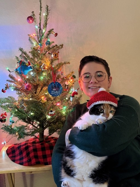 A person with glasses smiles while holding a cat wearing a red and white Santa hat, sitting beside a small decorated Christmas tree with colorful lights and ornaments.