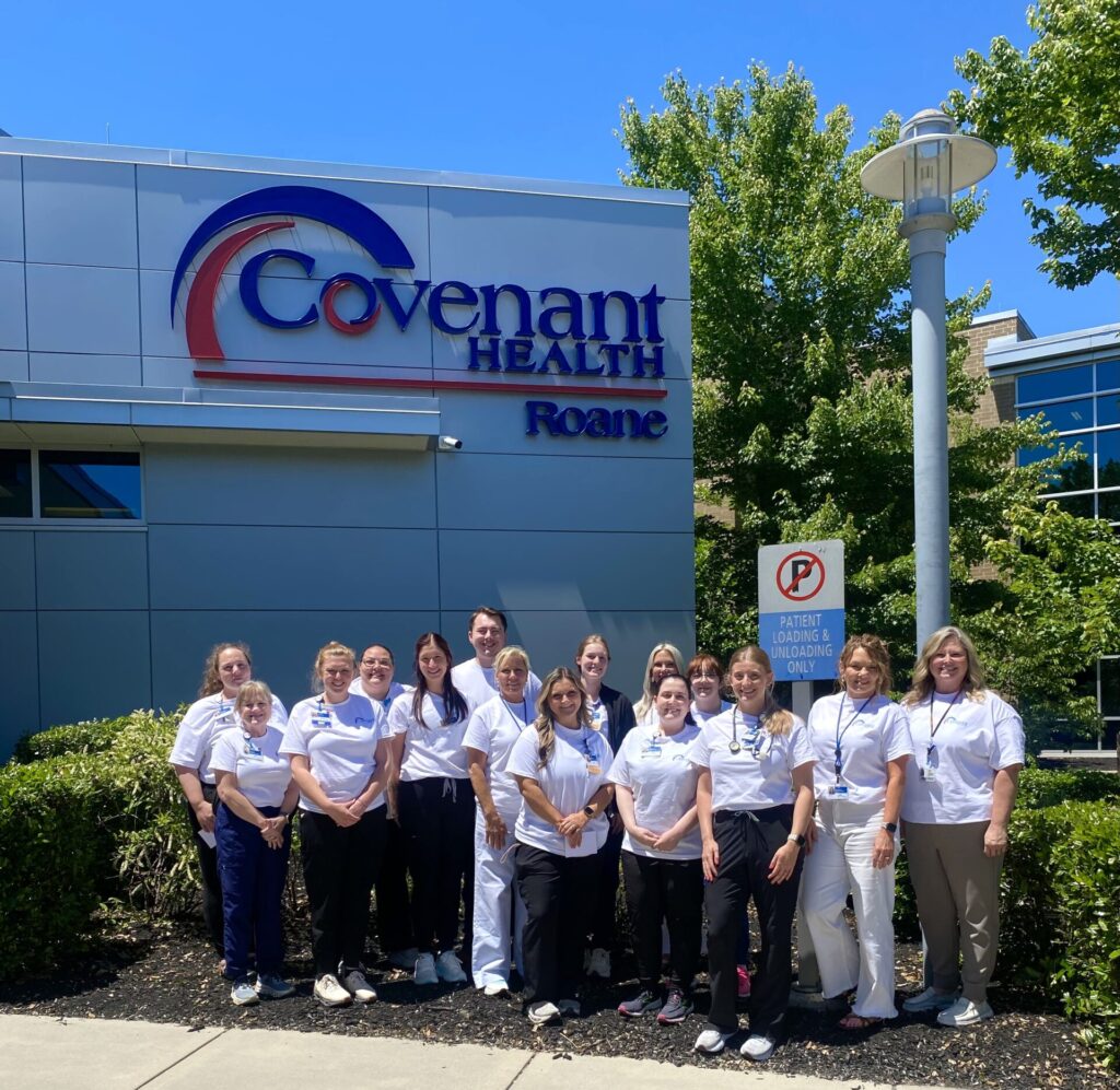 A group of people in white shirts stand smiling in front of a building with a sign that reads Covenant Health Roane on a sunny day, with trees and a no parking sign nearby.