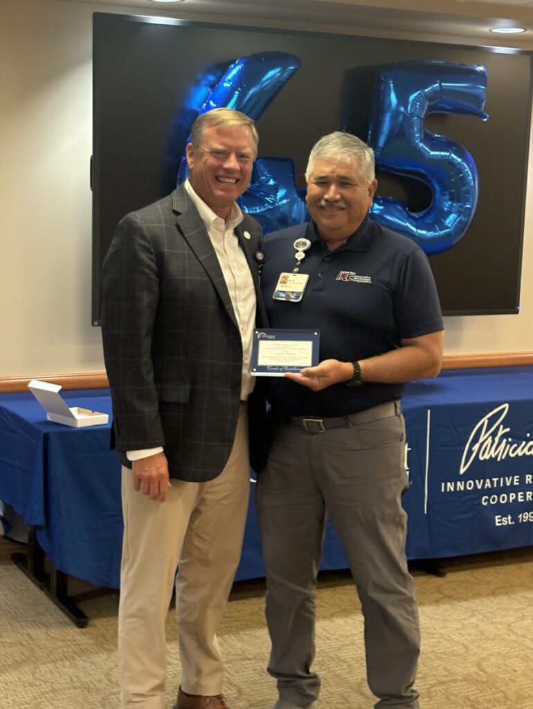 Two men smiling, one in a blazer and the other in a polo shirt holding a plaque. Behind them are large balloon numbers 65 and a blue table with a Patricia sign, suggesting a celebration or award event.