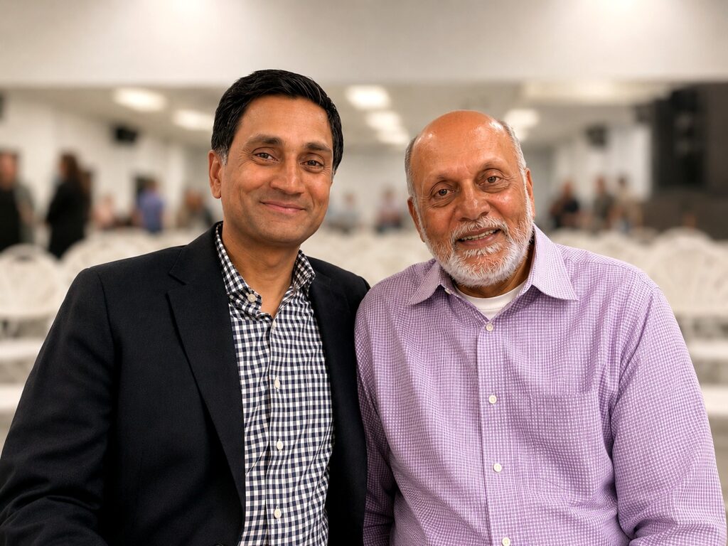 Two men sit closely together, smiling at the camera. One wears a dark blazer over a checkered shirt, and the other wears a light purple button-down shirt. The background is out of focus, with people and rows of white chairs visible.