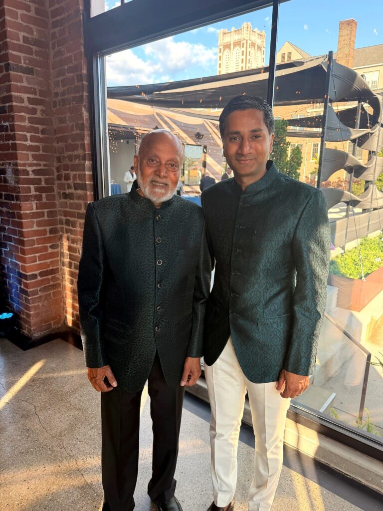 Two men stand indoors by large windows, both wearing dark green traditional jackets. The older man has white hair and a beard, while the younger man has short dark hair and wears white pants. Sunlight lights the scene.