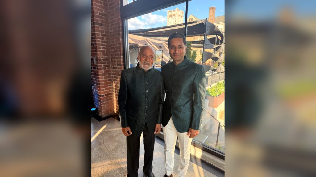 Two men stand side by side indoors, both wearing dark green traditional jackets. One man has white hair and a beard, while the other is younger with short hair and white pants. Sunlight streams through large windows behind them.