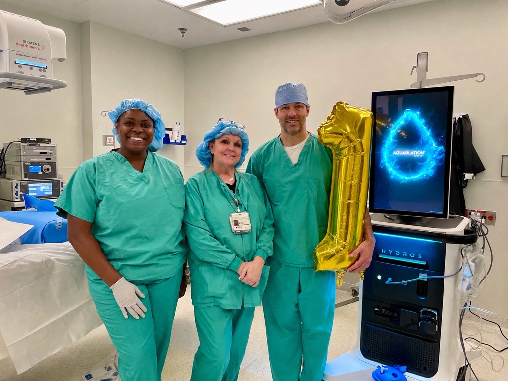 Three medical professionals in scrubs and hair covers stand in an operating room. One person holds a gold balloon shaped like the number 1. Medical equipment and a monitor displaying a blue logo are visible in the background.
